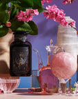 The Tea and Tarot to Go Gift Set from Magic Hour presents a purple-themed arrangement that includes a dark glass bottle, a clear glass pitcher filled with pink liquid, a rose quartz sphere, and various crystal formations surrounded by pink cherry blossoms, all set against a purple background to evoke the Tarot archetype.