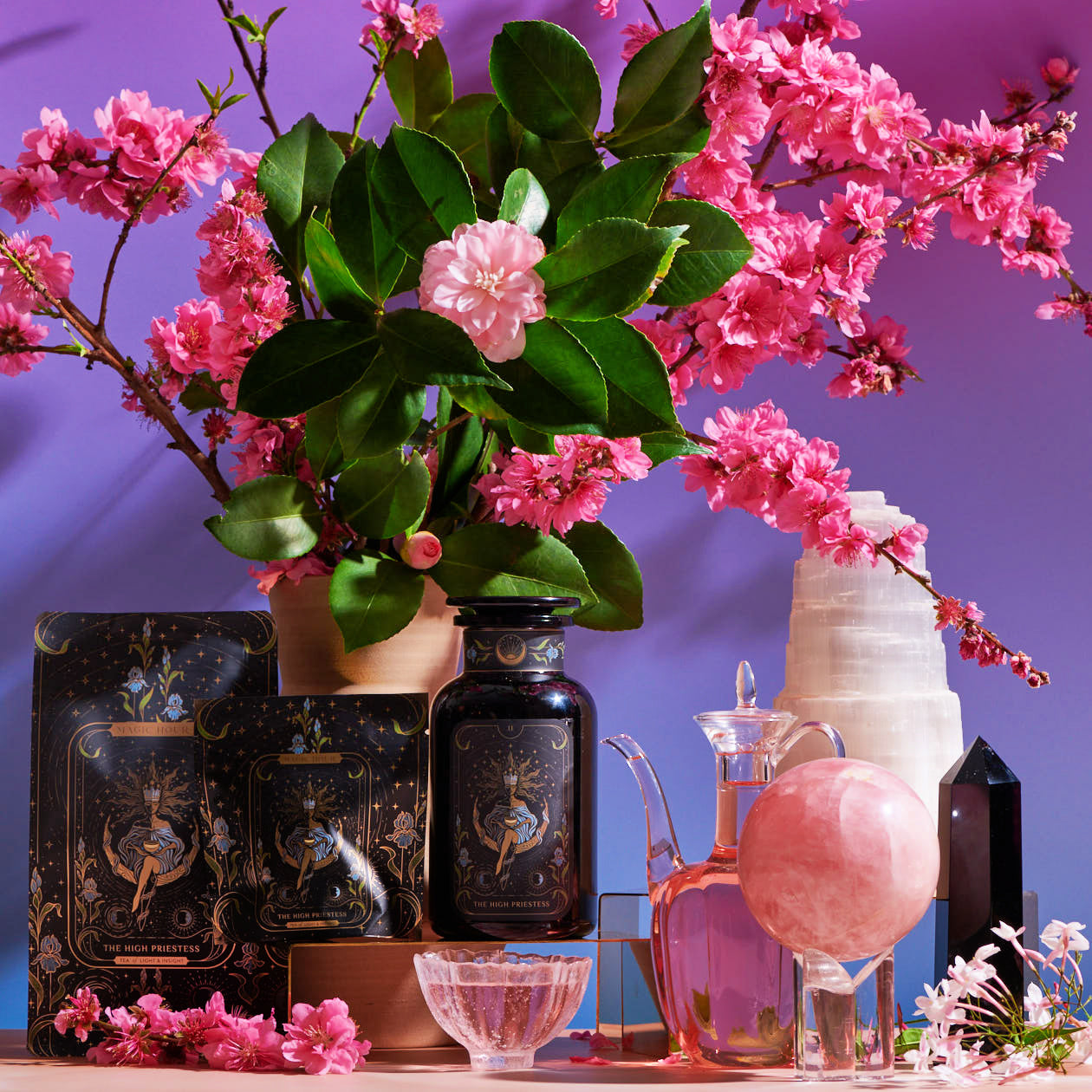 A decorative setup featuring The High Priestess Box skincare products with mystical-themed packaging, surrounded by pink cherry blossoms and green leaves. The display includes an ornate glass pitcher, a pink crystal sphere on a stand, a black crystal tower, and vintage glassware. A pastel background enhances the vibrant colors of The High Priestess Box.