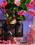 A decorative setup featuring The High Priestess Box skincare products with mystical-themed packaging, surrounded by pink cherry blossoms and green leaves. The display includes an ornate glass pitcher, a pink crystal sphere on a stand, a black crystal tower, and vintage glassware. A pastel background enhances the vibrant colors of The High Priestess Box.