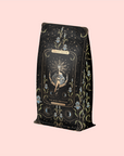 A decorative tea package with an ornate celestial and floral design features a central illustration of a seated woman. The label reads "Magic Hour" and "The High Priestess." This organic, caffeine-free herbal tea comes in a soft pink background. Product: The High Priestess Ginger-Mint-Cardamom Tea Refill Pouch.