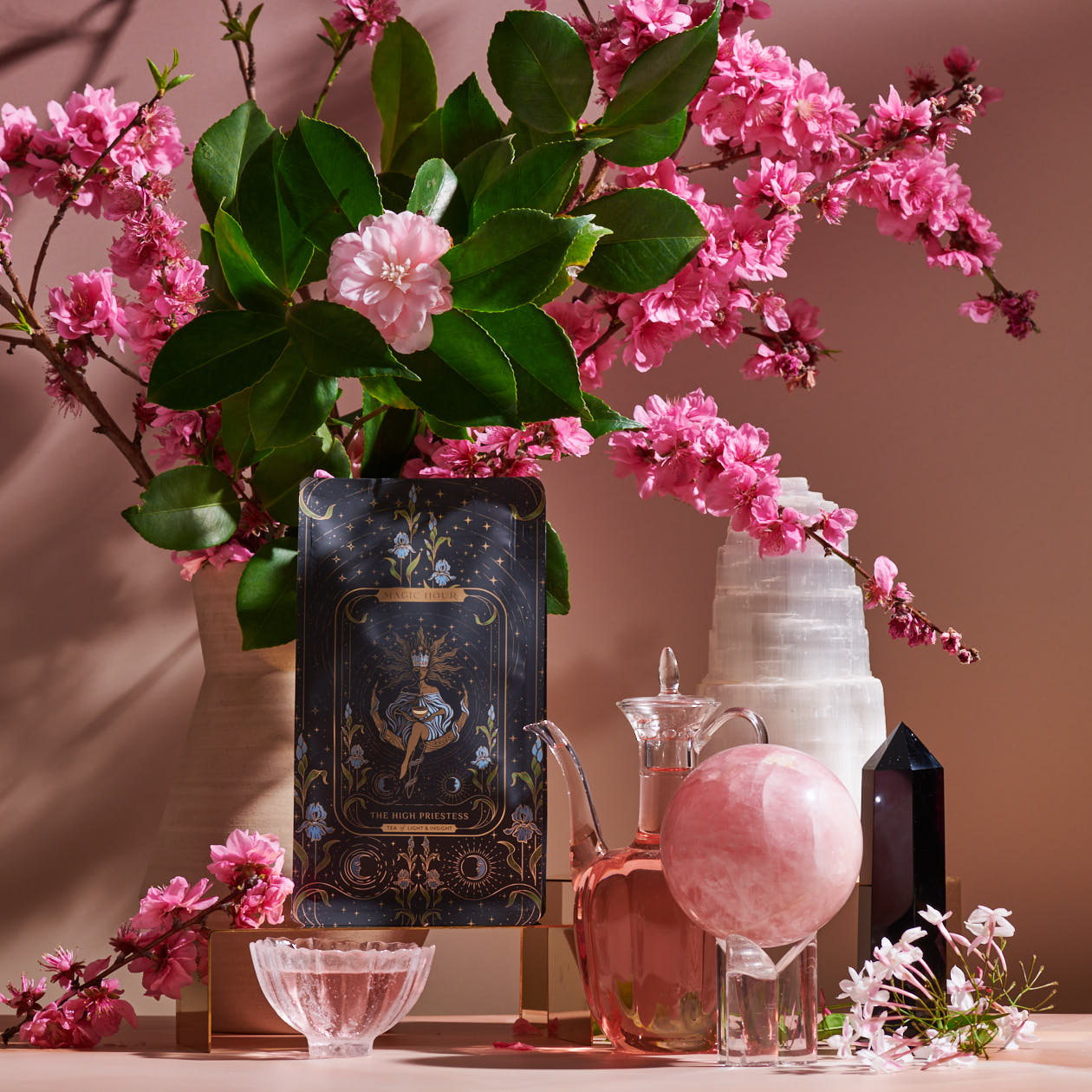 A still life scene with a vase of pink flowers and green leaves, a decorative tarot card from The High Priestess Box, a glass pitcher filled with pink liquid, a clear bowl, a rose quartz sphere, and both black and white crystals. The background is softly illuminated with a warm, pink hue.