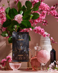 A still life scene with a vase of pink flowers and green leaves, a decorative tarot card from The High Priestess Box, a glass pitcher filled with pink liquid, a clear bowl, a rose quartz sphere, and both black and white crystals. The background is softly illuminated with a warm, pink hue.