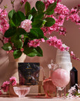 The High Priestess Box showcases a decorative arrangement with pink blossoms in a beige vase, a black card adorned with artwork, a glass carafe filled with pink liquid reminiscent of High Priestess Wisdom Tea, a crystal sphere, an elegant tall white crystal, and a striking black crystal obelisk—all set against a soothing pink background.