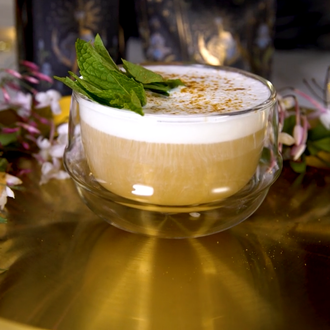 A frothy tea from The High Priestess Box, topped with mint leaves and a sprinkle of spices, sits in a double-walled glass cup on a reflective golden surface. Surrounding the cup are pink and white flowers, adding a touch of elegance to the presentation.