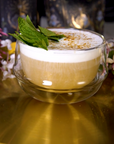 A glass cup filled with The High Priestess: Wisdom Tea for Powerful Dreams & Illuminated Insights by Magic Hour, garnished with fresh mint leaves on top, sits on a golden surface. In the background, there are floral elements and ornate black and gold decorative items.