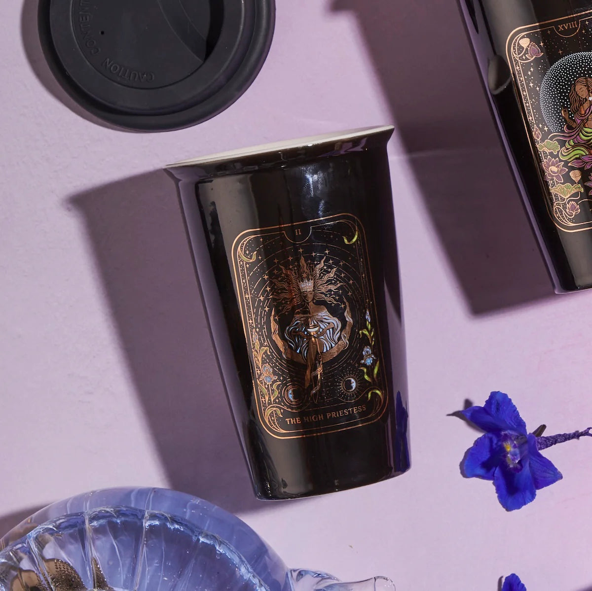 A black cup adorned with intricate golden designs, featuring an image reminiscent of 'The High Priestess' tarot archetype, rests on a lavender surface. Nearby sit another cup with a similar motif, blue flowers, and a black lid. Ideal for the Magic Hour Tea and Tarot to Go Gift Set or savoring your personalized tea blend.