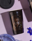 A black cup adorned with intricate golden designs, featuring an image reminiscent of 'The High Priestess' tarot archetype, rests on a lavender surface. Nearby sit another cup with a similar motif, blue flowers, and a black lid. Ideal for the Magic Hour Tea and Tarot to Go Gift Set or savoring your personalized tea blend.