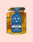 The Honey Comb Hex Jar - Acacia by Savannah Bee Company is displayed against a pink background. This 12 oz (340 g) jar features a gold lid, blue label with a honeybee design, and contains premium pure honey and natural acacia raw honeycomb.