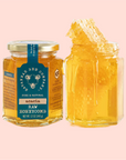 Two glass jars are showcased: one with a teal label reading "Honey Comb Hex Jar - Acacia," and the other open, showing honey with edible honeycomb inside, all set against a light pink background.