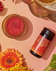 An arrangement features a black and red bottle of Hydrate Tea Boost with gold accents, a small dish of red powder, and a variety of flowers including sunflowers and orange gerbera daisies. A decorative tray with assorted items is partially visible in the background.