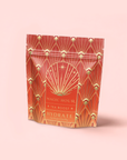 A red and gold resealable pouch labeled "Hydrate Tea Boost" with an art deco pattern and starburst design stands upright on a light pink background, emphasizing its hydration benefits from nutrient-rich botanicals.