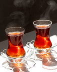 Two Turkish tea glasses filled with hot Organic Iyerpadi Estate Black Tea rest on glass saucers, steam swirling upward as sunlight highlights reflections and shadows across the white surface beneath them.