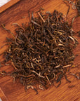 A close-up view of a pile of Jasmine Yin Hao Green Tea by Magic Hour on a wooden surface. The tea leaves are loosely spread out, showcasing their dark, twisted, and curled textures. The wooden background adds a natural, rustic feel to the image.