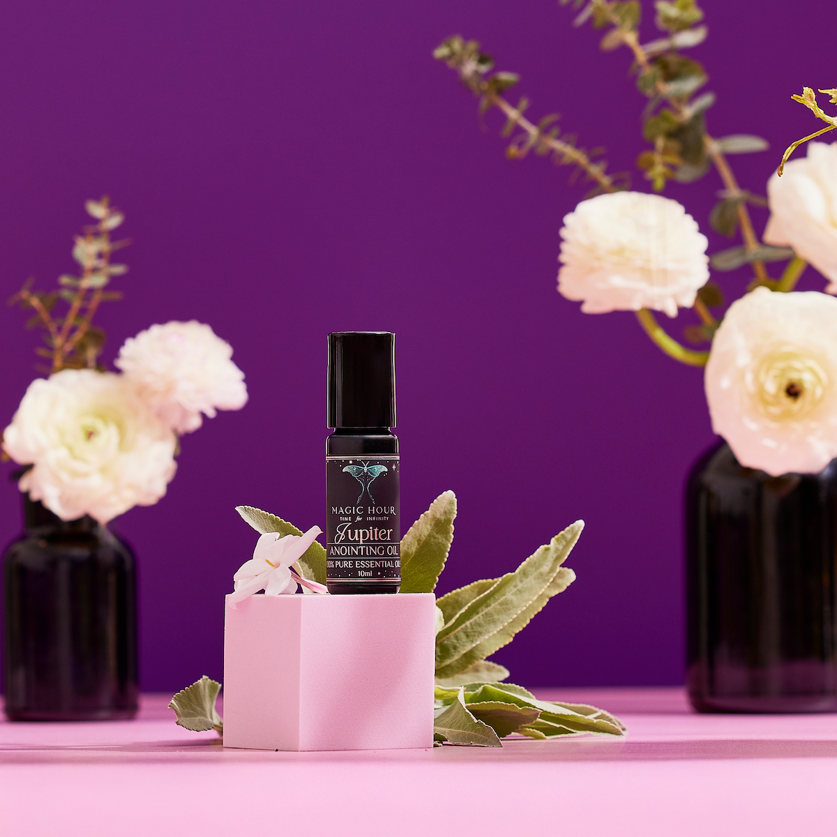 A small black bottle labeled "Planetary Perfume Oil" sits on a pink cube with green leaves and a white flower, set against a purple background with white flowers in black vases.