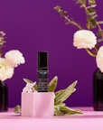 A small black bottle labeled "Planetary Perfume Oil" sits on a pink cube with green leaves and a white flower, set against a purple background with white flowers in black vases.