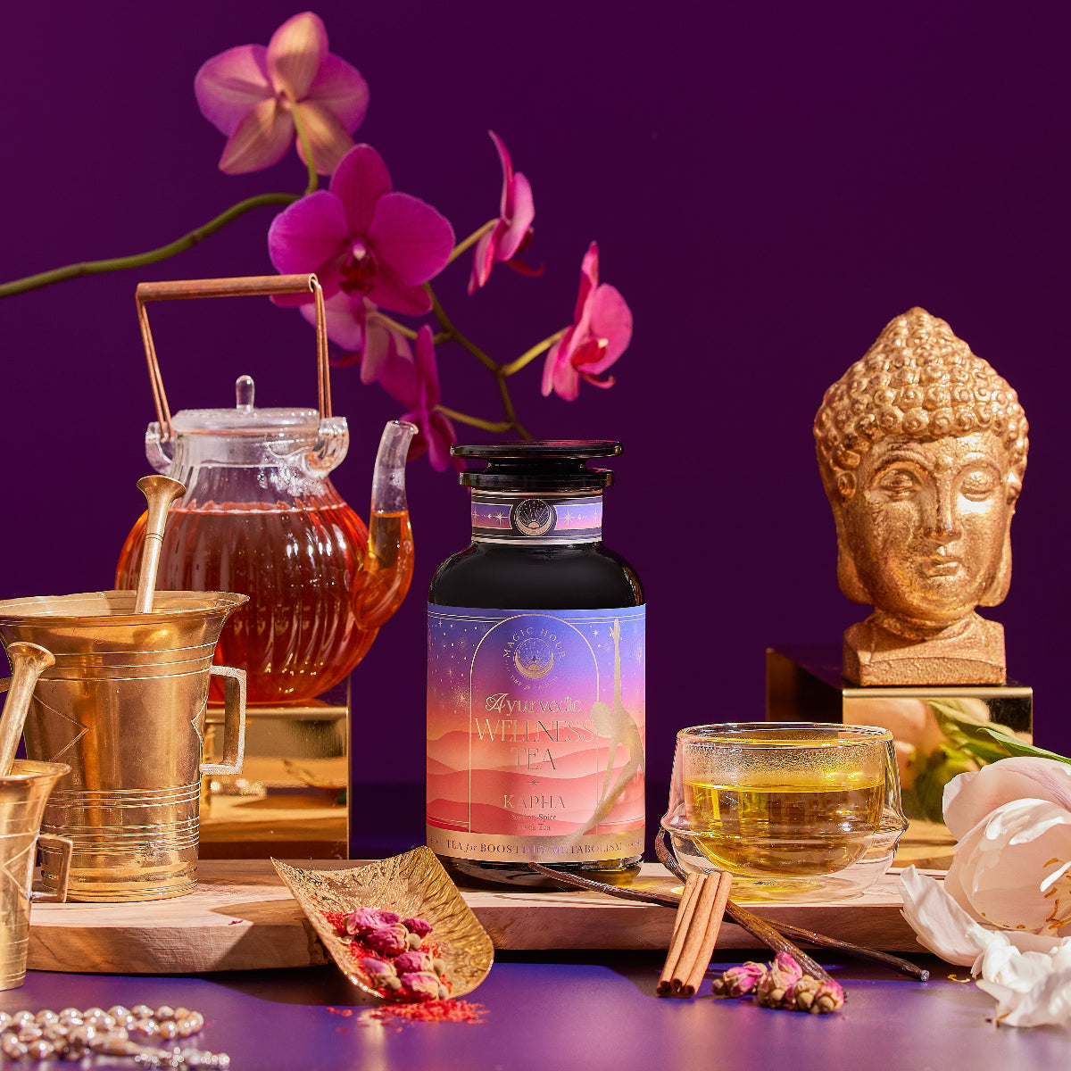 The Kapha Herbal bottle sits on a wooden tray with a glass teapot, teacup, golden mortar and pestle, pink orchids, a golden Buddha statue, and various spices, all set against a deep purple background.