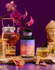 The Kapha Herbal bottle sits on a wooden tray with a glass teapot, teacup, golden mortar and pestle, pink orchids, a golden Buddha statue, and various spices, all set against a deep purple background.