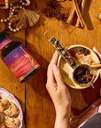 A person holds a mug of chai with star anise and cinnamon on a wooden table surrounded by Kapha Herbal supplement, cookies, sliced apples, and festive decor.