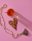 A cup of Balancing Dosha Bundle adaptogenic tea rests on a pink surface beside a heart of loose leaves and petals, accented by a beaded tassel necklace surrounding the arrangement.