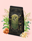A black and gold tea bag labeled “Kettle Corn Kiss: Sweet & Nutty Toasted Rice Green Tea with Caramel & Vanilla” is surrounded by illustrated plants, caramel pieces, and popcorn on a light pink background—evoking the warm, toasty notes of toasted rice green tea complemented by the sweetness of caramel and vanilla.