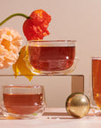 Two Kinto Double-Walled Teacups filled with tea sit beside a metal tea infuser. Two colorful poppy flowers hang over the scene, and a third glass of tea rests in the background—all arranged on a light pink surface.