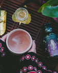 A person in a patterned sleeve holds a mug of tea above a wooden surface, near a jar of Aphrodite's Alchemy: Damiana & Rose White Tea, a gold incense holder with burning incense, and a glass of tea—evoking calming rituals.