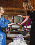 Two women smile as they exchange a Branded Gift Box, surrounded by flowers, tea jars, and crystals on a cozy, ornate wooden bench.