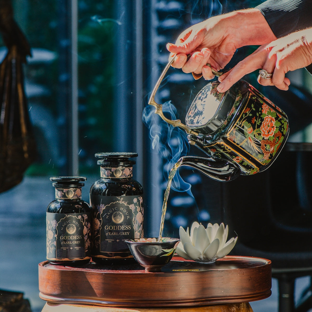 A person pours organic tea from an ornate teapot into a small cup. The table features two black jars labeled "Goddess of Earl: Lady Luck Oriental Beauty Oolong Tea" by Magic Hour. A white lotus-shaped ornament is also on the table. Steam rises from the cup, suggesting the tea is hot, adding to the serene magic hour of tea time.
