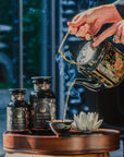 A person pours organic tea from an ornate teapot into a small cup. The table features two black jars labeled "Goddess of Earl: Lady Luck Oriental Beauty Oolong Tea" by Magic Hour. A white lotus-shaped ornament is also on the table. Steam rises from the cup, suggesting the tea is hot, adding to the serene magic hour of tea time.