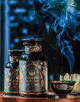 Two dark glass jars labeled "Goddess of Earl: Lady Luck Oriental Beauty Oolong Tea" from Magic Hour are placed on a wooden surface. A small bowl releases smoke, contributing to the serene ambiance. The background is softly blurred, accentuating the aromatic setting of this organic loose leaf tea captured in the image.