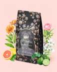A beautifully floral-decorated tea bag labeled "Goddess of Earl: Lady Luck Oriental Beauty Oolong Tea" rests amid vibrant flowers, fresh grapefruit, and bergamot on a pale pink background, evoking the delicate aroma of an Oriental Beauty Oolong blend.