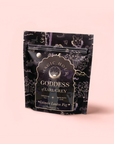 A black and gold pouch of Magic Hour’s Goddess of Earl: Lavender London Fog—Tea for Blooming Clarity & Calm Moods stands upright on a pale pink backdrop, featuring botanical illustrations and labeled as ceremonial bergamot lavender tea.