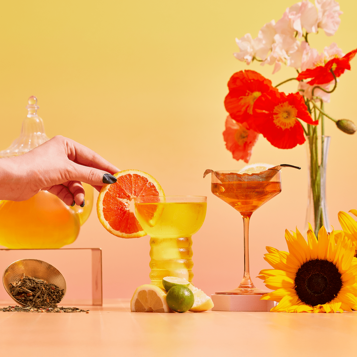 A hand adds a grapefruit slice to a yellow cocktail glass. Next to it is Lemon Lime Green Tea: Ceremonial Soda Pop Tea with Sugar "Destroyer", loose tea, lemon, lime, and flowers on a gradient yellow-to-orange backdrop.
