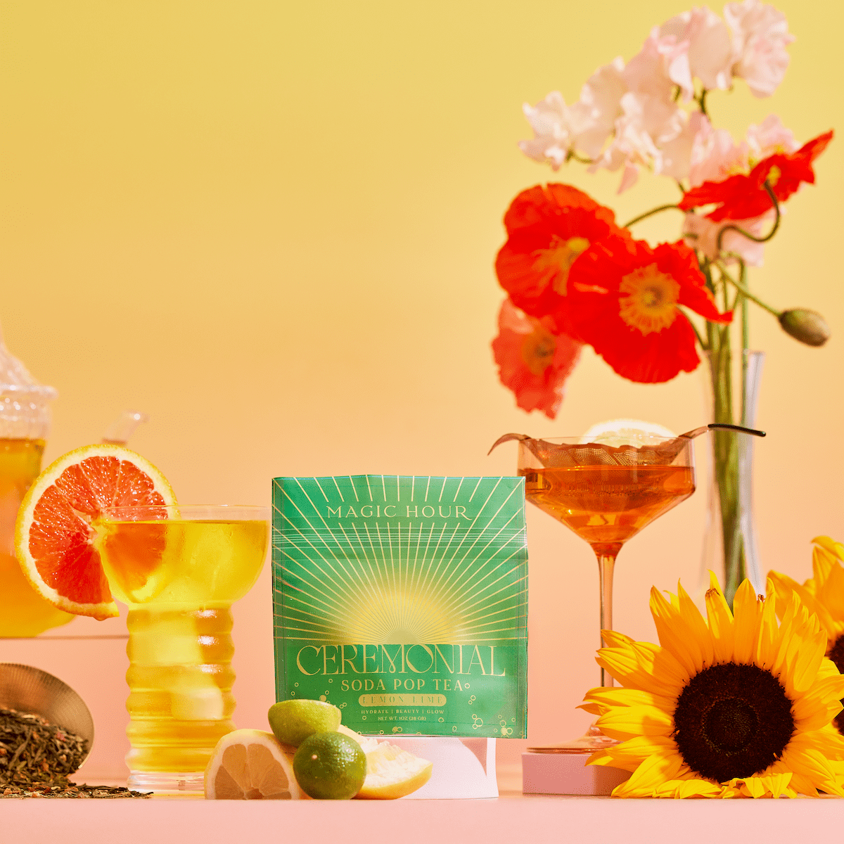 A Lemon Lime Green Tea: Ceremonial Soda Pop Tea with Sugar "Destroyer" box sits beside a glass of green tea with grapefruit, a cocktail, sliced lemon and lime, a sunflower, and red and white flowers on a warm yellow gradient background.