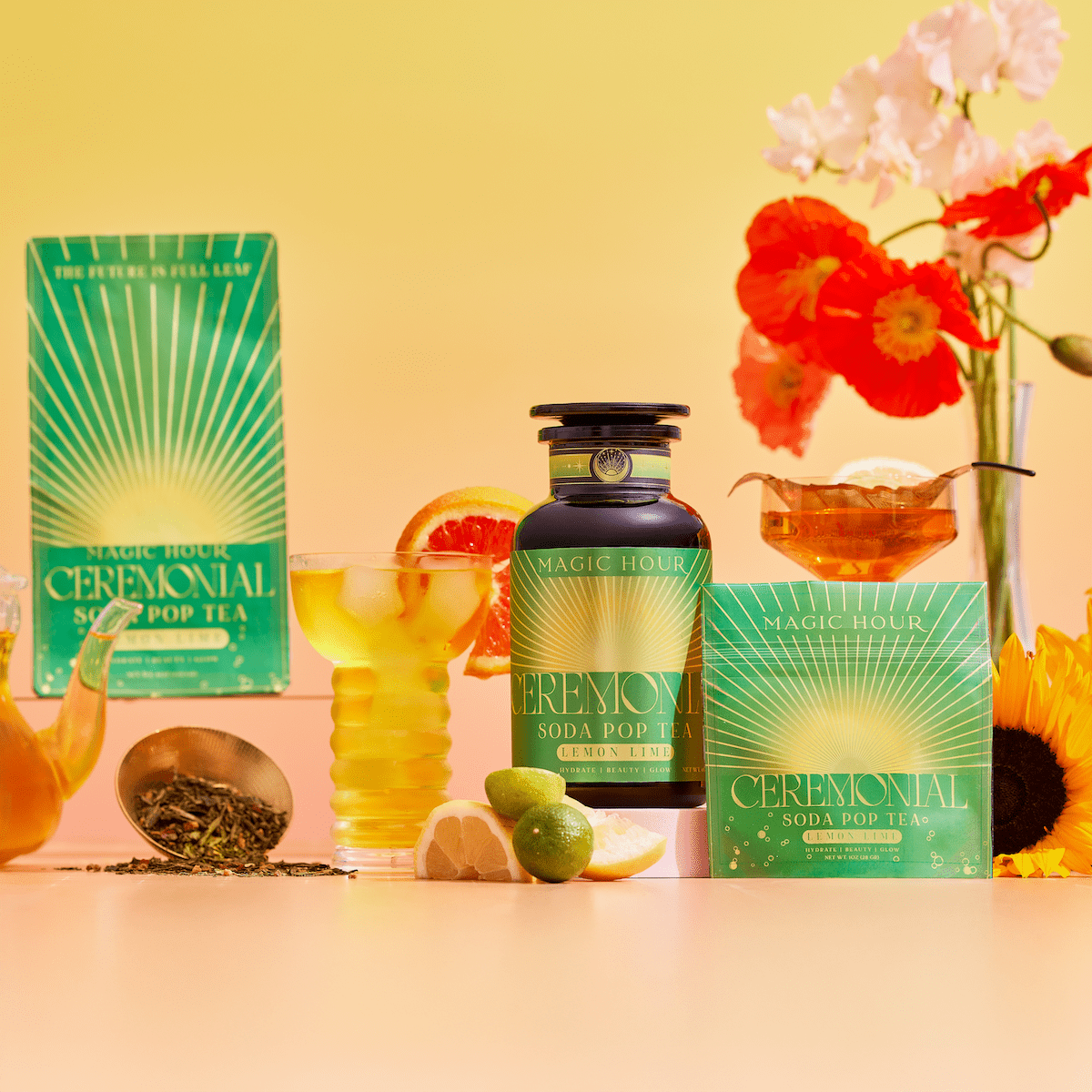 A vibrant display of Lemon Lime Green Tea: Ceremonial Soda Pop Tea with Sugar "Destroyer"—featuring its tin, packaging, iced tea with citrus, fresh flowers, loose green tea leaves, and fruit—all set against a warm yellow-orange background.