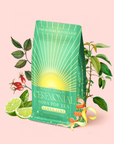 A green bag of Lemon Lime Green Tea: Ceremonial Soda Pop Tea is displayed on a light pink background, surrounded by fresh lemon and lime slices, rose hips, and tea leaves. Accented with golden rays and elegant typography, this vibrant blend harmoniously combines the zesty brightness of lemon tea with premium green tea leaves and a unique touch of gymnema sylvestre.