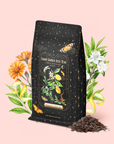 A black, starry tea bag labeled "Lemon Meringue Puerh Black Iced Tea" stands with loose tea leaves, surrounded by illustrated flowers, lemons, and greenery on a light pink background. This lemon puerh black iced tea is known for supporting healthy digestion, offering a delightful and probiotic tea experience.