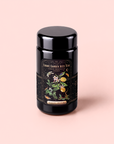 A sleek black tea canister adorned with a vibrant, botanical label that reads “Cosmic Garden Iced Teas” and “Magic Hour,” showcases playful illustrations of fruits and flowers on a soft pink background. This elegant packaging highlights the Lemon Meringue Puerh Black Iced Tea, an invigorating blend crafted for those seeking a refreshing and revitalizing weight loss tea experience.