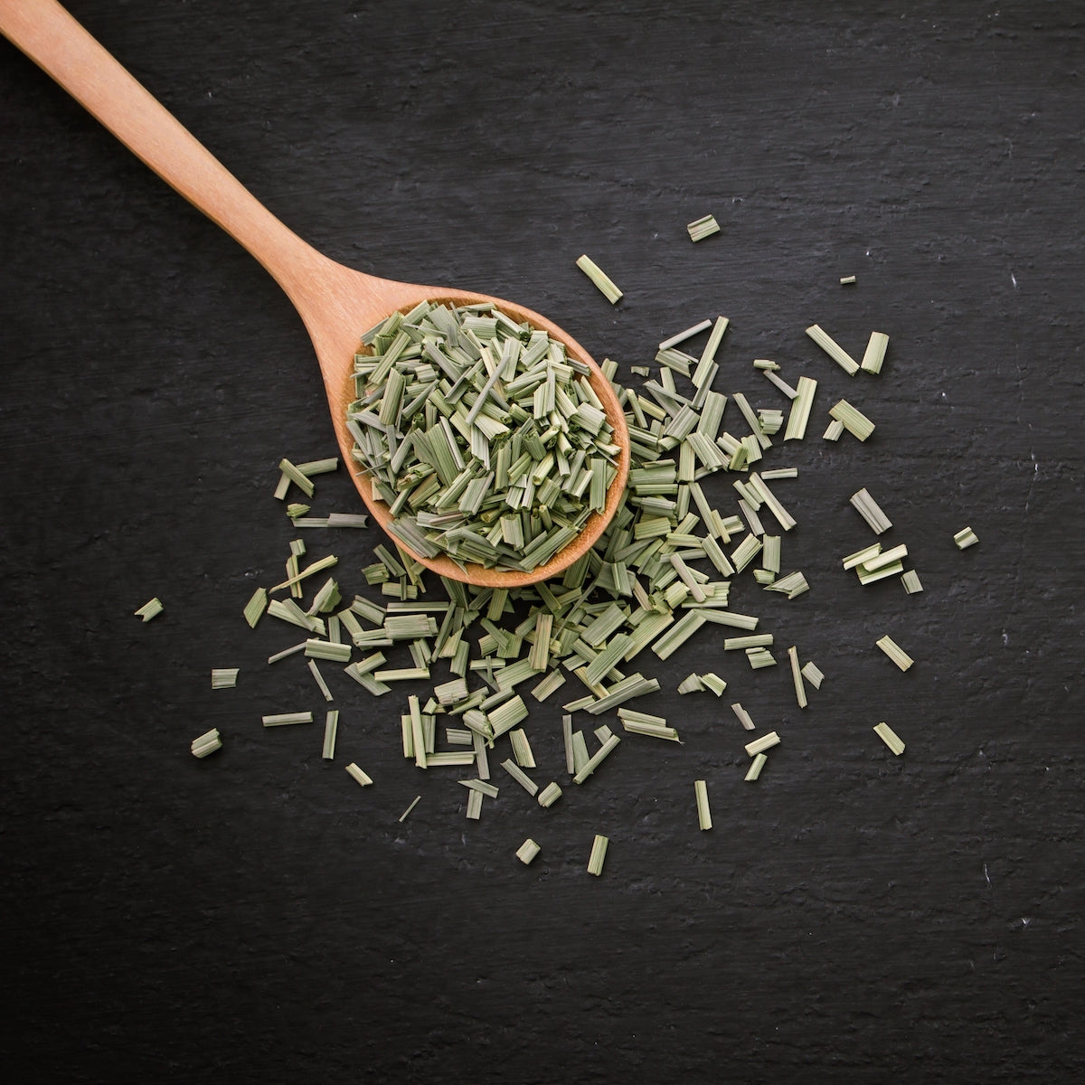 A wooden spoon filled with dried, green, chopped herbs rests on a dark, textured surface. Some of the herbs are scattered around the spoon, creating a casual, organic arrangement.