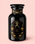 A black glass bottle features a decorative label with a golden figure holding a crescent moon, surrounded by flowers and stars. The label displays “Leo: Queen of Courage Tea of the Sun” and “Magic Hour” on a pale pink background—hinting at mystical blends within, such as Yunnan Black Tea Pearls or the invigorating touch of Lion’s Mane.