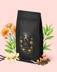 The Leo: Queen of Courage Tea of the Sun features a black tea bag with floral gold art and a dancing woman, surrounded by illustrated flowers, green leaves, Yunnan Black Tea leaves, vanilla beans, orange peels, and peppercorns on soft pink.