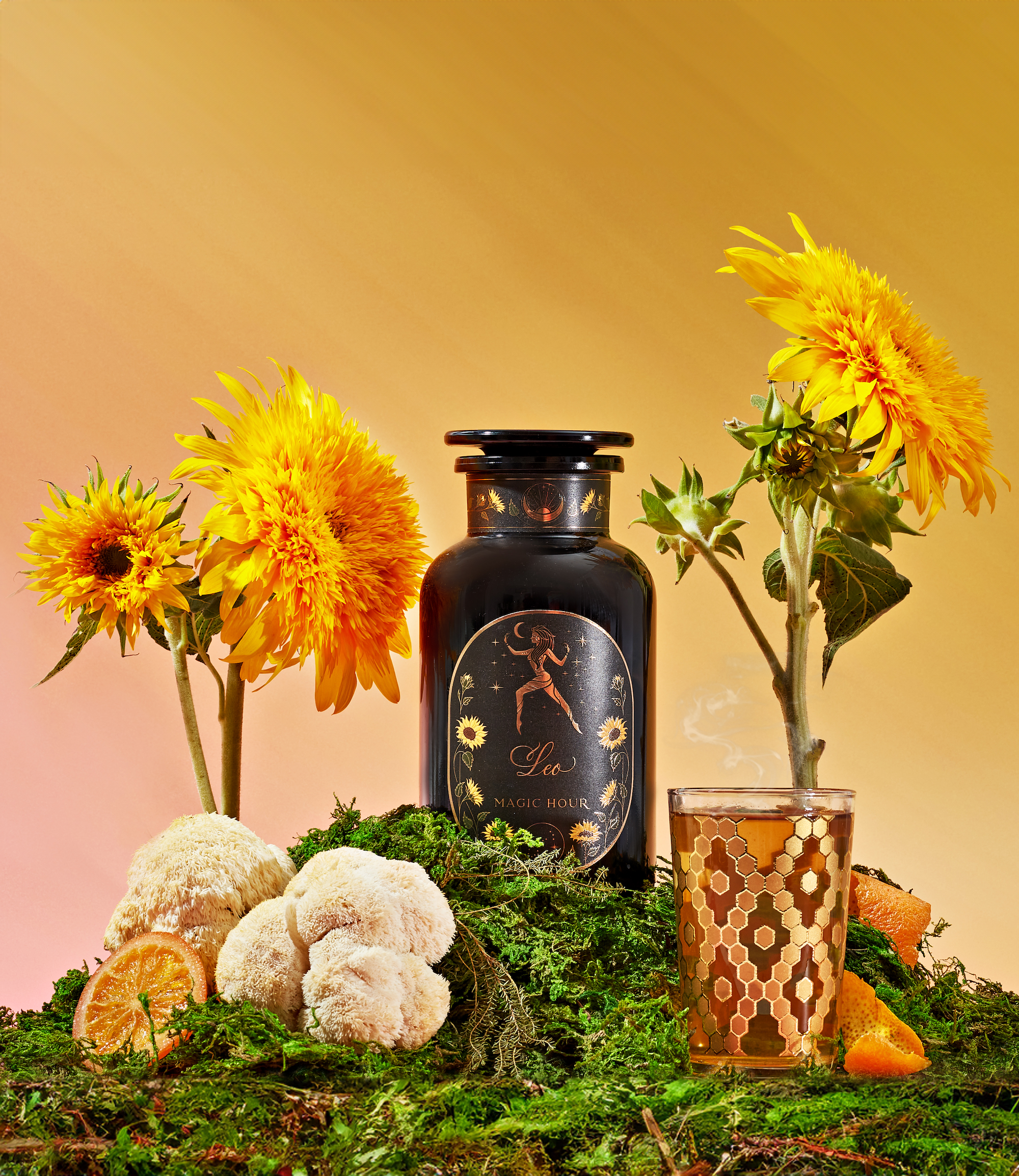 A still life composition featuring a black jar labeled "Leo Magic Hour" amidst vibrant yellow sunflowers and moss. A glass of tea with a decorative pattern is placed beside the jar, complemented by orange slices, white mushrooms, and green herbs.
