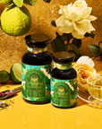 Two ornate green tea jars labeled "London Fog Green" sit amid fresh limes, roses, and tea leaves. A clear glass teacup filled with tea rests on a vibrant yellow surface, exuding floral complexity against a gold textured background.