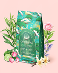 A green tea package labeled "London Fog Green" is surrounded by botanicals, including roses, lavender, bergamot, lime, and white flowers, set on a light pink background. The package features birds and floral patterns.