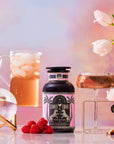 A bottle labeled "Summer Fridays™: Rose All Day," an alcohol-free rosé alternative, sits on a table with raspberries, a pink drink in a glass, iced beverage pitcher, crystal ball, and white tulips against a pastel background.
