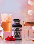 A bottle labeled "Summer Fridays™: Rose All Day," an alcohol-free rosé alternative, sits on a table with raspberries, a pink drink in a glass, iced beverage pitcher, crystal ball, and white tulips against a pastel background.