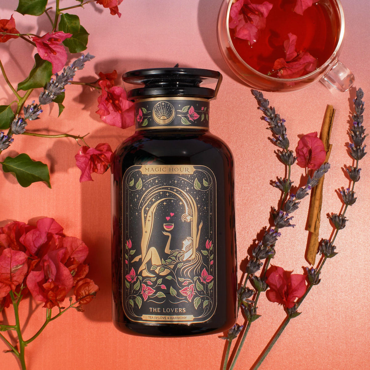 A jar of "The Lovers Box" tea blend rests on a pink background, surrounded by sprigs of lavender, pink flowers, and a cinnamon stick. A glass cup of tea is partially visible in the top right corner. The jar features an illustration of a romantic scene.
