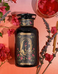 A jar of "The Lovers Box" tea blend rests on a pink background, surrounded by sprigs of lavender, pink flowers, and a cinnamon stick. A glass cup of tea is partially visible in the top right corner. The jar features an illustration of a romantic scene.
