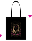 A black tote bag features an illustration titled "Magic Hour: The Lovers," depicting a romantic scene with a couple surrounded by roses and stars. Three floating pink hearts accent the right side of the tote bag, making it an ideal companion to The Lovers Box tea blend.