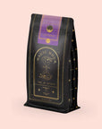 The "Lucid Dreams" Herbal Tea Refill Pouch is a black, gold, and purple bag of Magic Hour turmeric tulsi tea with an elegant hourglass and celestial design. The label reads "Lucid Dreams" and "Time for Infinity." This caffeine-free blend stands against a light beige background.